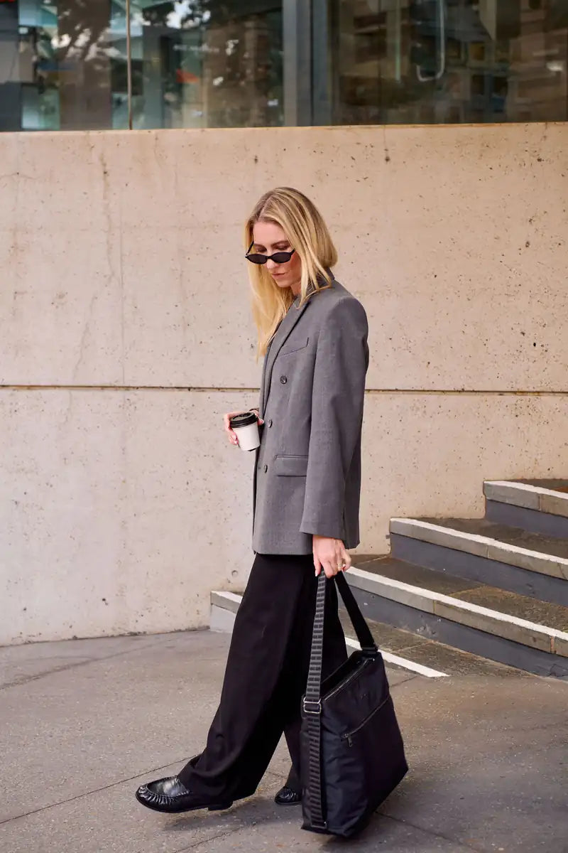 A woman walking outdoors wearing an oversized gray blazer, wide-leg black pants, black shoes, and black sunglasses, carrying a black tote bag and holding a takeaway coffee cup.