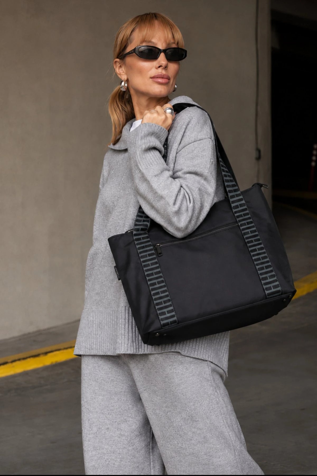 A woman in a gray knit lounge set and black sunglasses carries a large black tote bag with gray striped handles in an indoor parking structure.