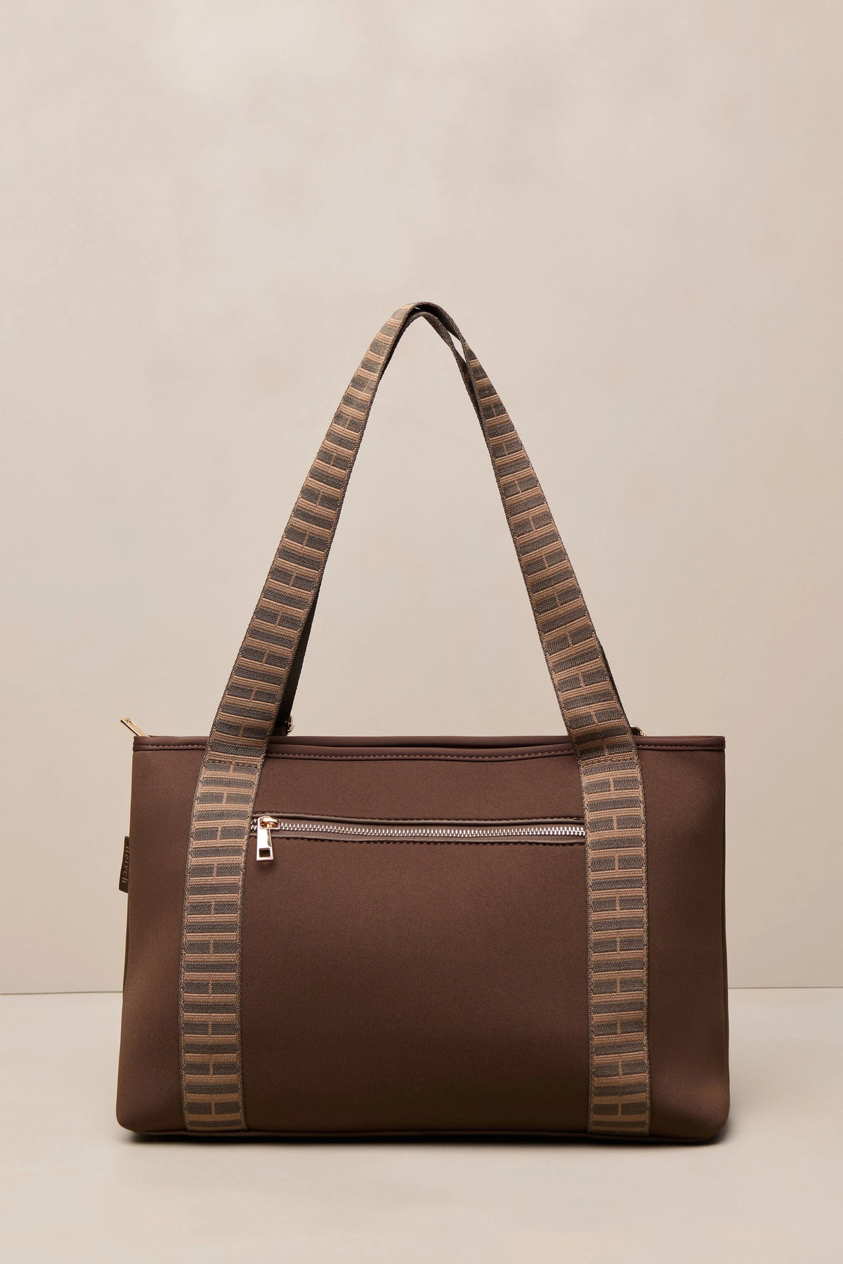 A brown chocolate tote bag with patterned shoulder straps, featuring a front zippered pocket and gold-tone hardware, set against a neutral beige background.