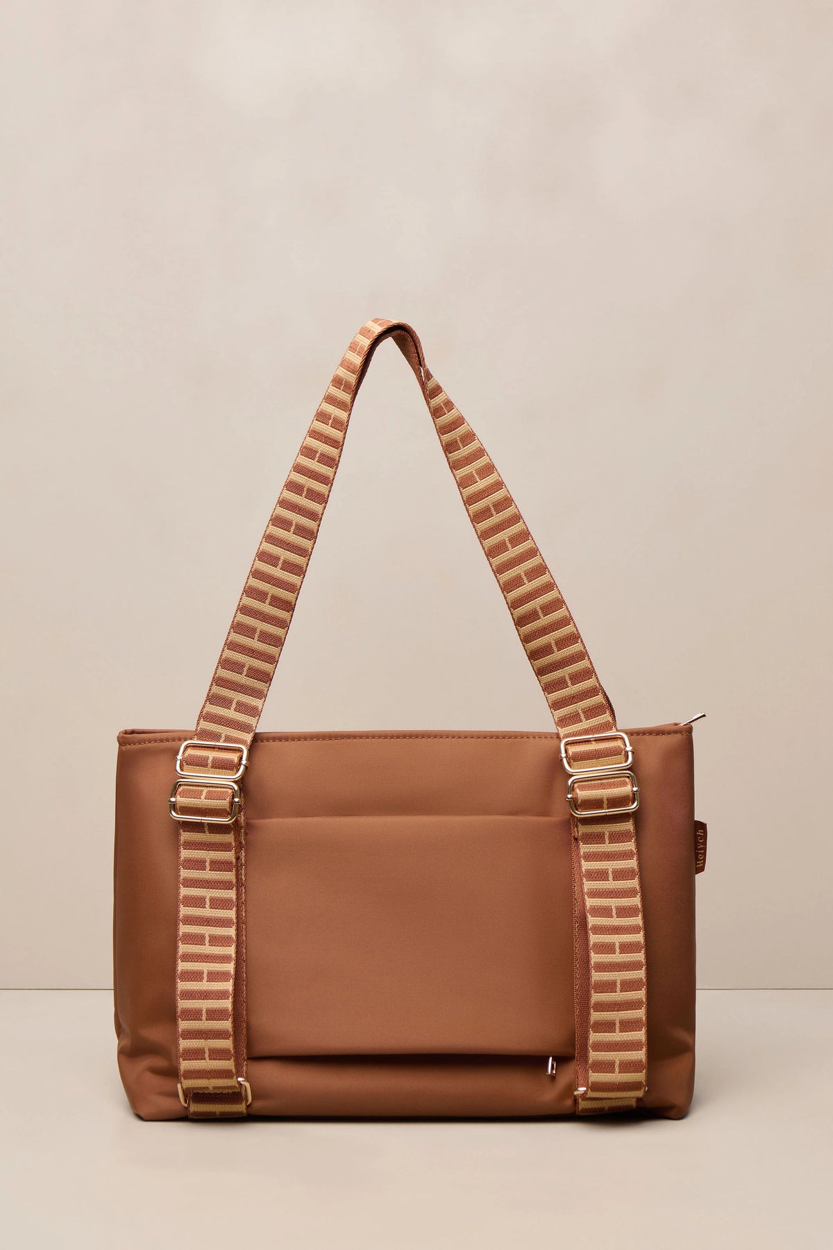 A cognac nylon tote bag with a matching brown woven shoulder strap featuring beige stripe details and gold-tone metal hardware, shown against a neutral beige background.