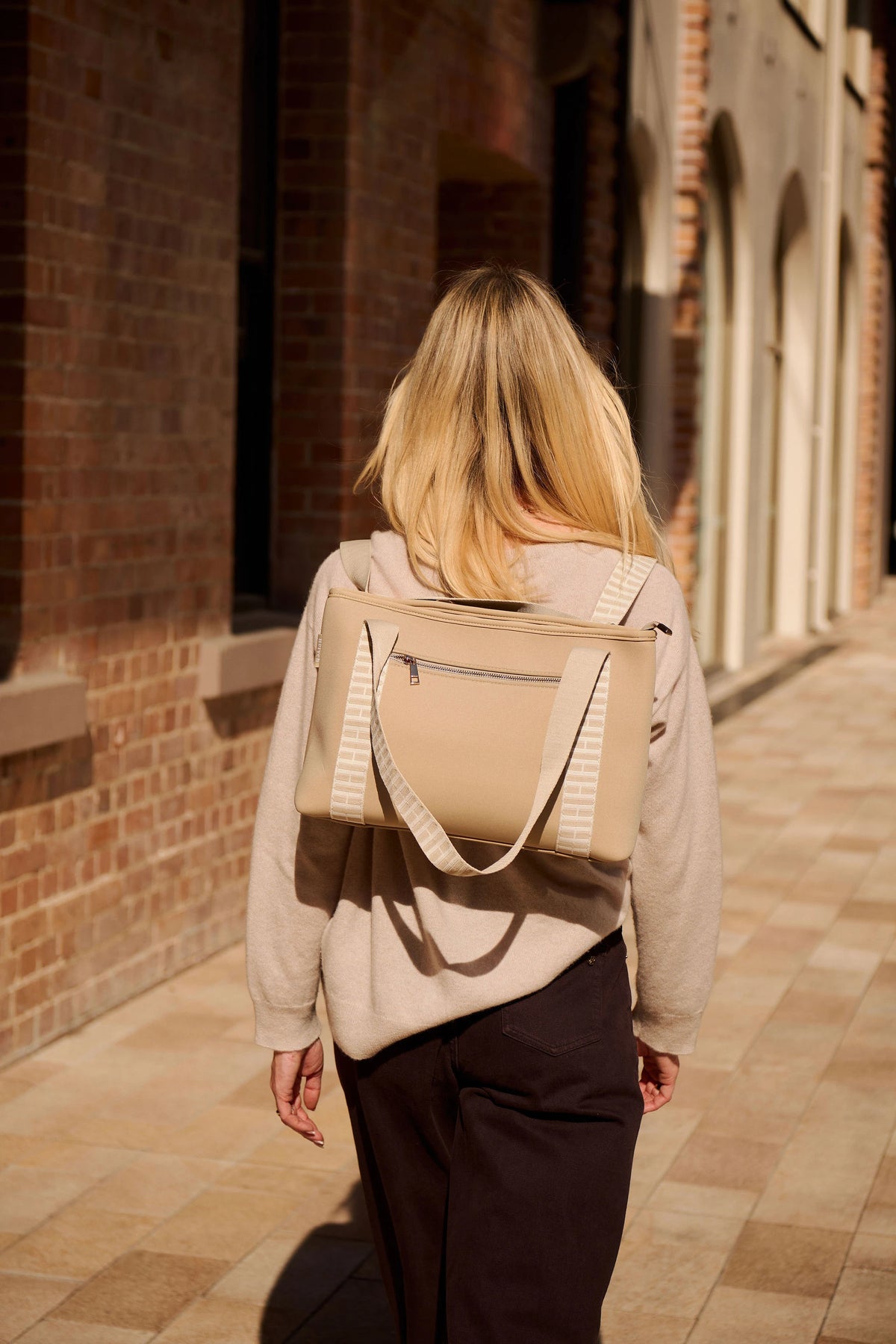 A woman viewed from behind walking down a sunlit brick walkway, wearing a taupe beige tote bag over her shoulders as a backpack. The bag has a zippered top and striped fabric side straps, styled with a beige hoodie and dark pants.