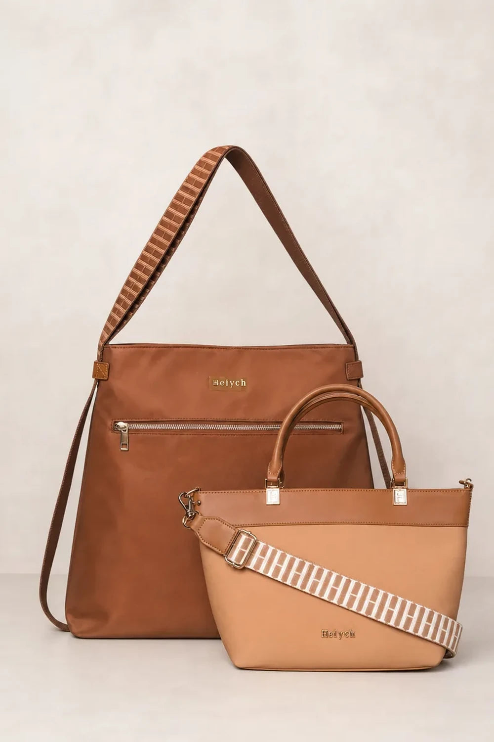 Two cognac tan handbags, one larger brown shoulder bag and one smaller tan tote bag, both featuring striped woven shoulder straps and silver-tone hardware, set against a neutral light beige background.