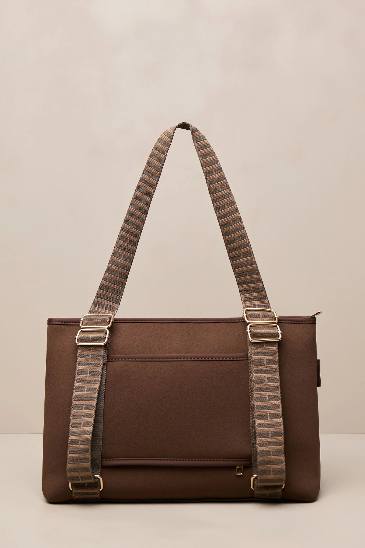 A brown chocolate shoulder bag with wide, patterned brown and beige woven straps and gold-tone hardware, photographed against a neutral beige background.