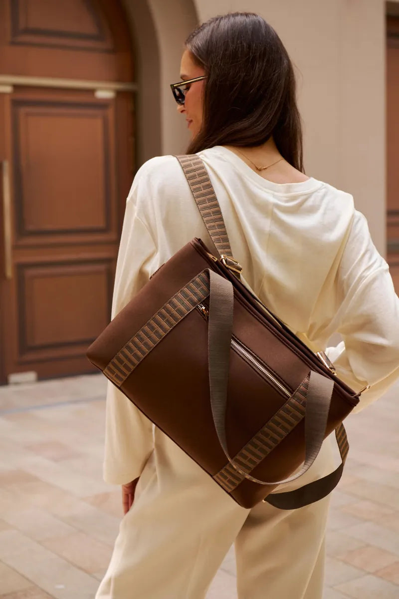 A woman wearing a cream-colored outfit and sunglasses carries a brown chocolate shoulder bag with patterned trim, photographed from behind in an outdoor setting.