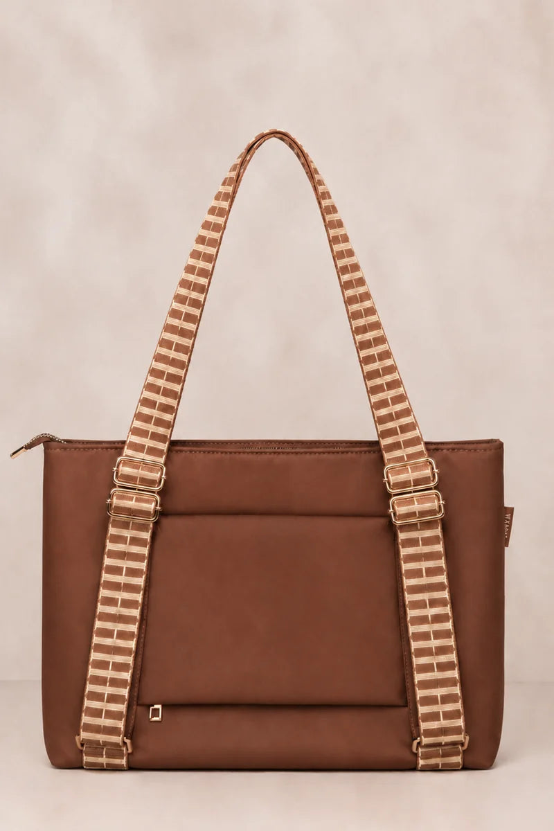 A brown cognac tote bag with wide, patterned brown and beige woven shoulder straps, photographed against a neutral beige background.