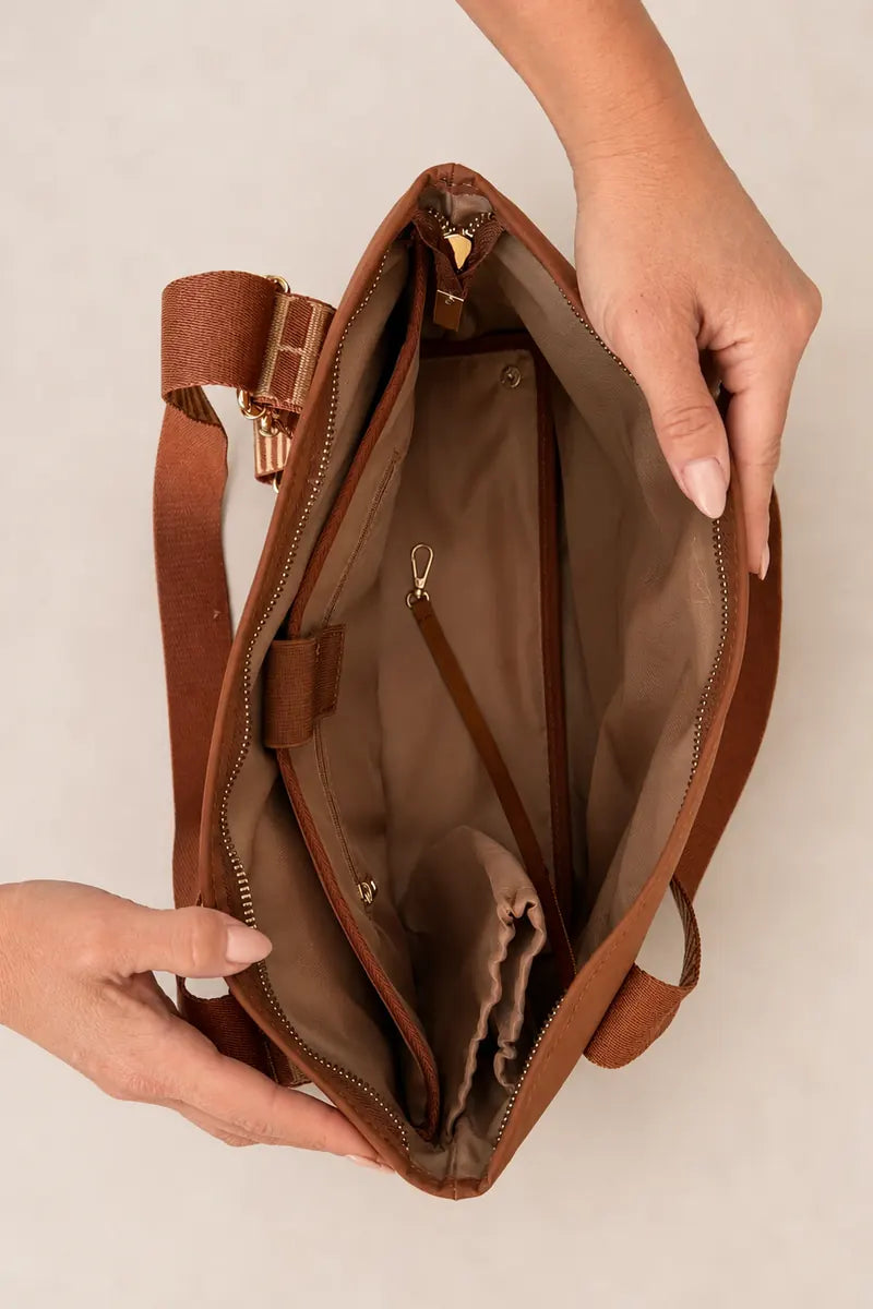 A brown cognac shoulder bag shown open from above, held by three hands to display its interior compartments, including a zippered pocket and a drawstring bag inside. The bag has a matching brown leather strap and gold-tone hardware.