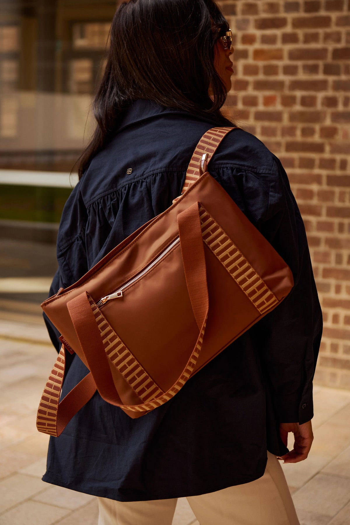 A woman wearing a navy blue jacket and beige pants is shown from behind, carrying a brown crossbody bag with patterned straps over her shoulder. The bag has a cognac brown body with beige and brown striped patterned straps and a silver zipper.
