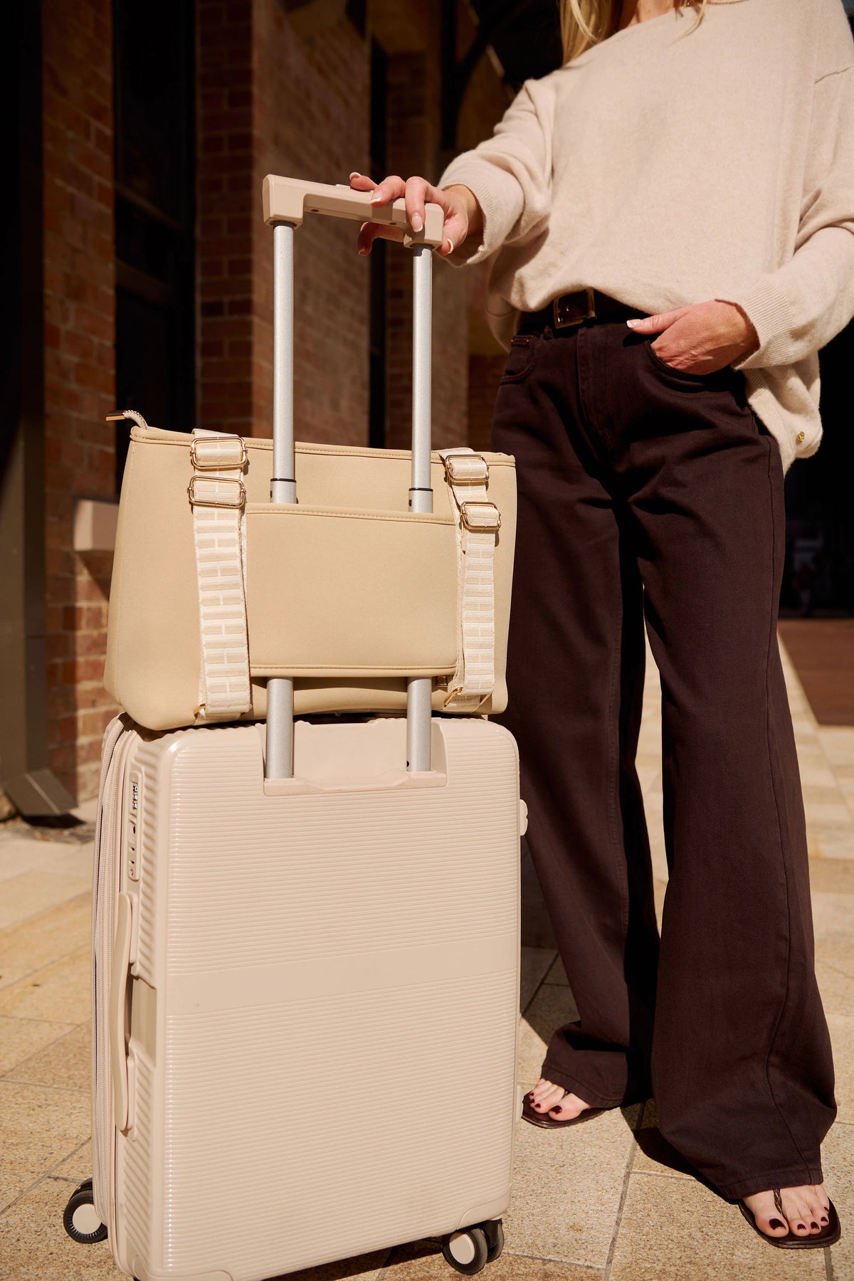 A woman in a cream sweater and dark brown wide-leg pants stands beside a beige hard-shell rolling suitcase with a matching taupe beige tote bag stacked on top, in an outdoor urban setting at night.