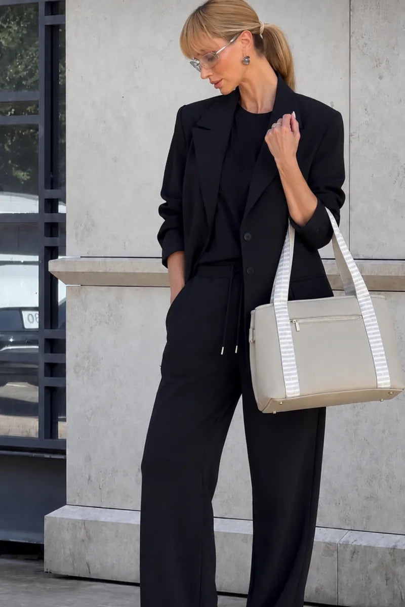 A woman wearing a tailored suit and holding a large cream-colored taupe tote bag with white striped handles, standing outdoors against a light gray stone wall.