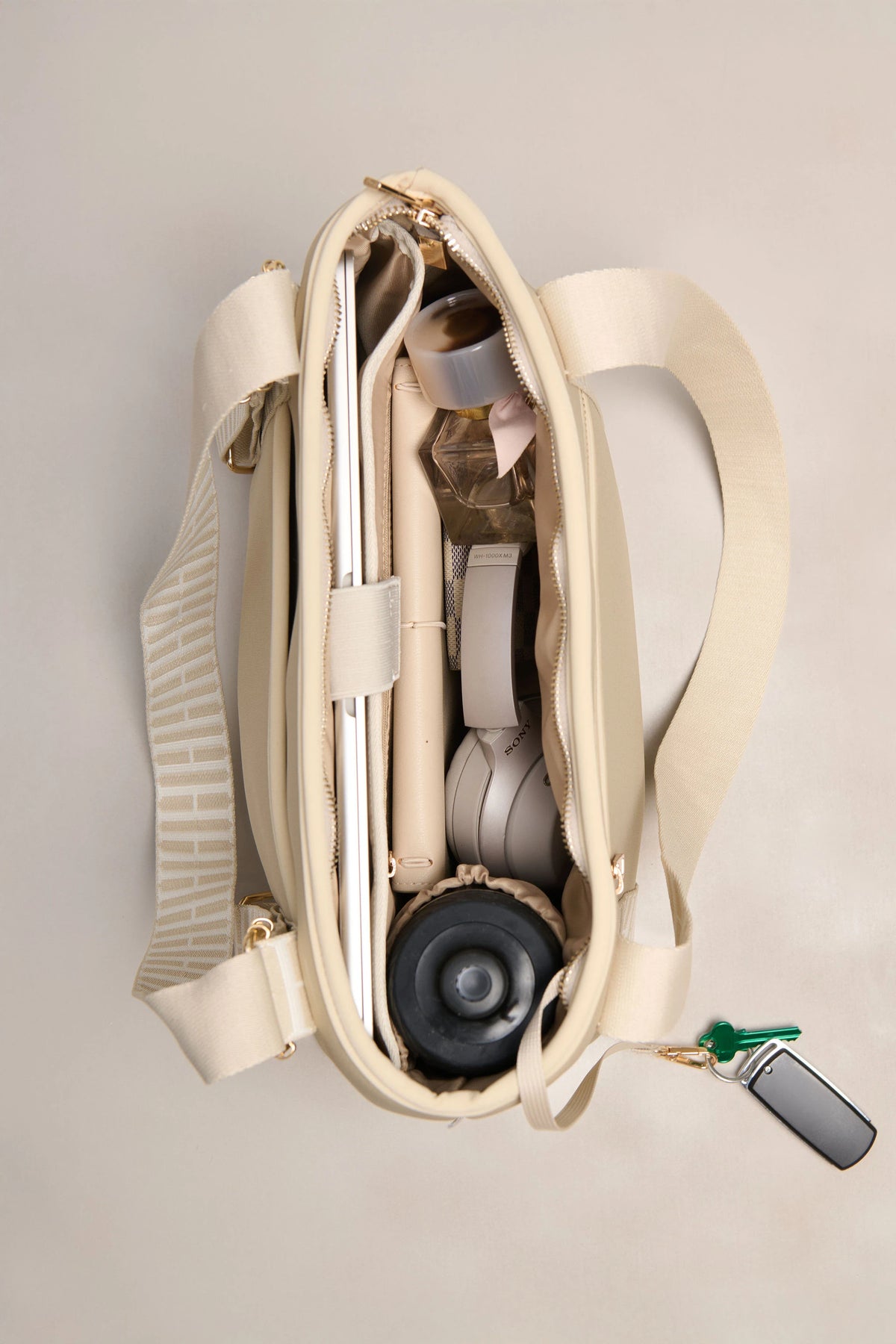 An open taupe-colored shoulder bag with a wide strap, shown from a top-down view on a light beige background. The bag is packed with various items, including a silver laptop, a pink pen, a white cord, and a drink bottle, with a small key fob hanging from the side zipper.