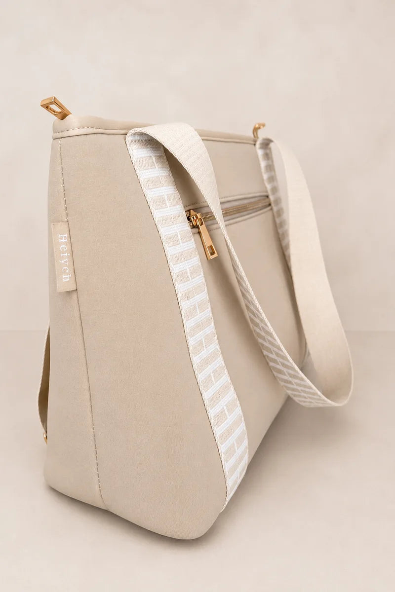 A structured taupe beige tote bag with a wide, adjustable shoulder strap featuring a white and beige geometric pattern, gold-tone hardware, and a small vertical brand tag on the side.