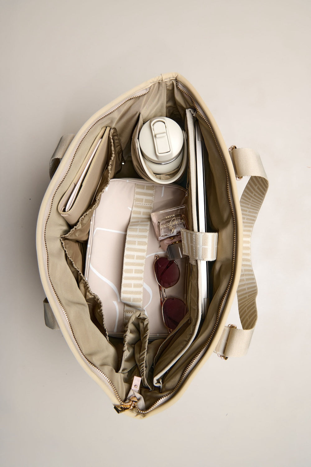 Open taupe tote bag with items including a white mug, sunglasses, and a book on a light gray background